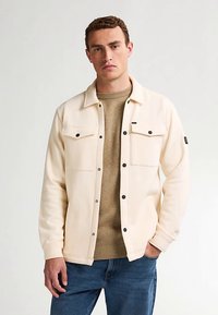 Kermanvärinen takki mustilla napeilla ja kahdella rintataskulla, päällä teksturoitu beige neule, yhdistettynä sinisiin farkkuihin.