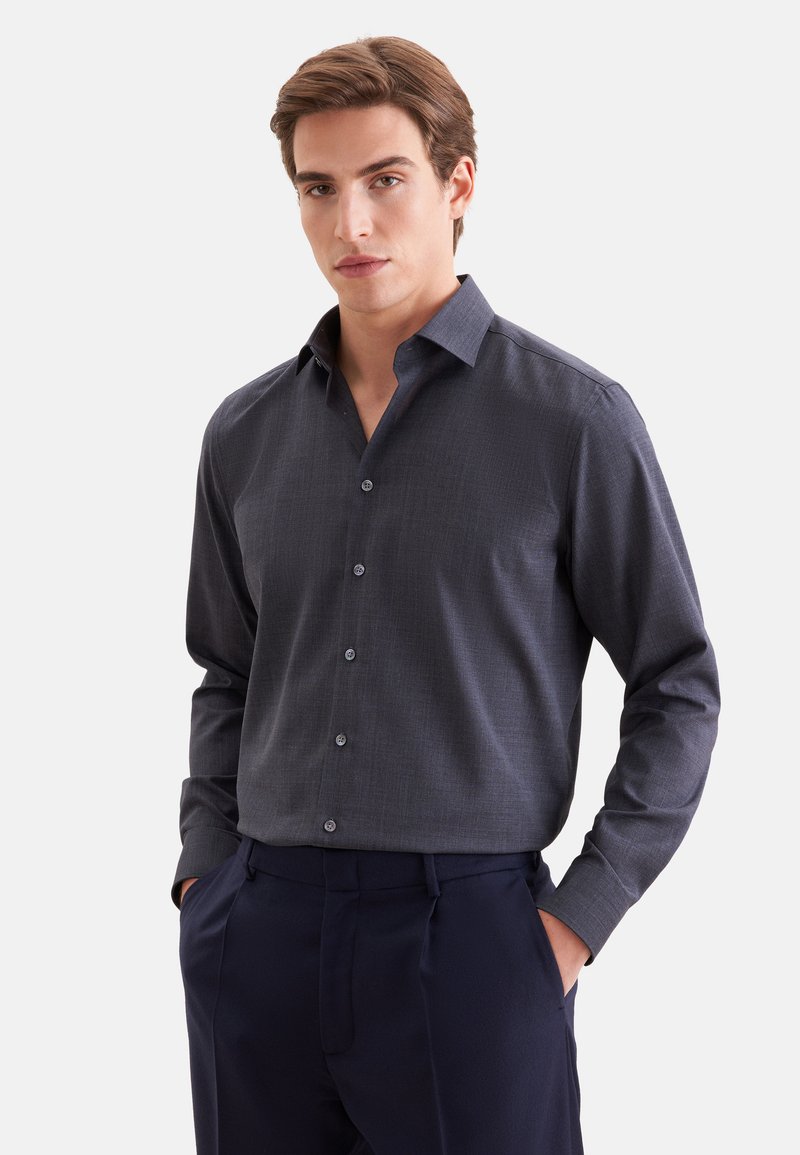 Jeune homme aux cheveux châtain clair portant une chemise boutonnée gris foncé et un pantalon foncé, debout les mains dans les poches devant un fond uni.