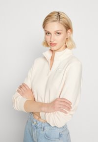 Svetloružový polovičný zips mikina z fleecu s vypchanými manžetami a malou vyšitou logom na hrudi, skombinovaná s džínsami s vysokým pásom v modrej farbe.