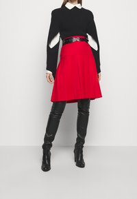 Schwarzer Strickpullover mit weißem Kragen, roter plissierter Rock mit schwarzem Gürtel und schwarze Leder-Stiefel bis zur Oberschenkel.