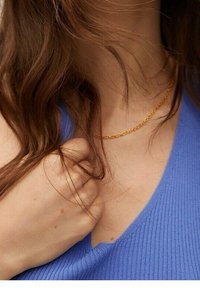 Złoty naszyjnik na szyi, charakteryzujący się delikatnym splatanym wzorem. Tło to niebieska materiałowa tkanina w prążki.