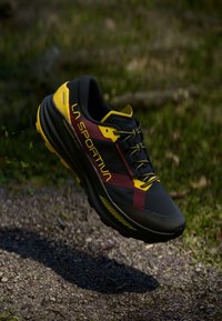 Melna un dzeltena taku skriešanas kurpe ar sarkaniem akcentiem un LA SPORTIVA apzīmogojumu, kas karājas virs grants taka ar zaļumiem fonā.