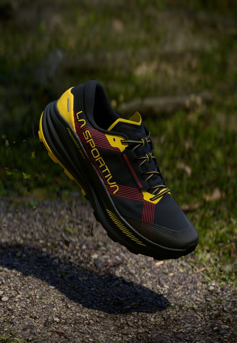 Melna un dzeltena taku skriešanas kurpe ar sarkaniem akcentiem un LA SPORTIVA apzīmogojumu, kas karājas virs grants taka ar zaļumiem fonā.