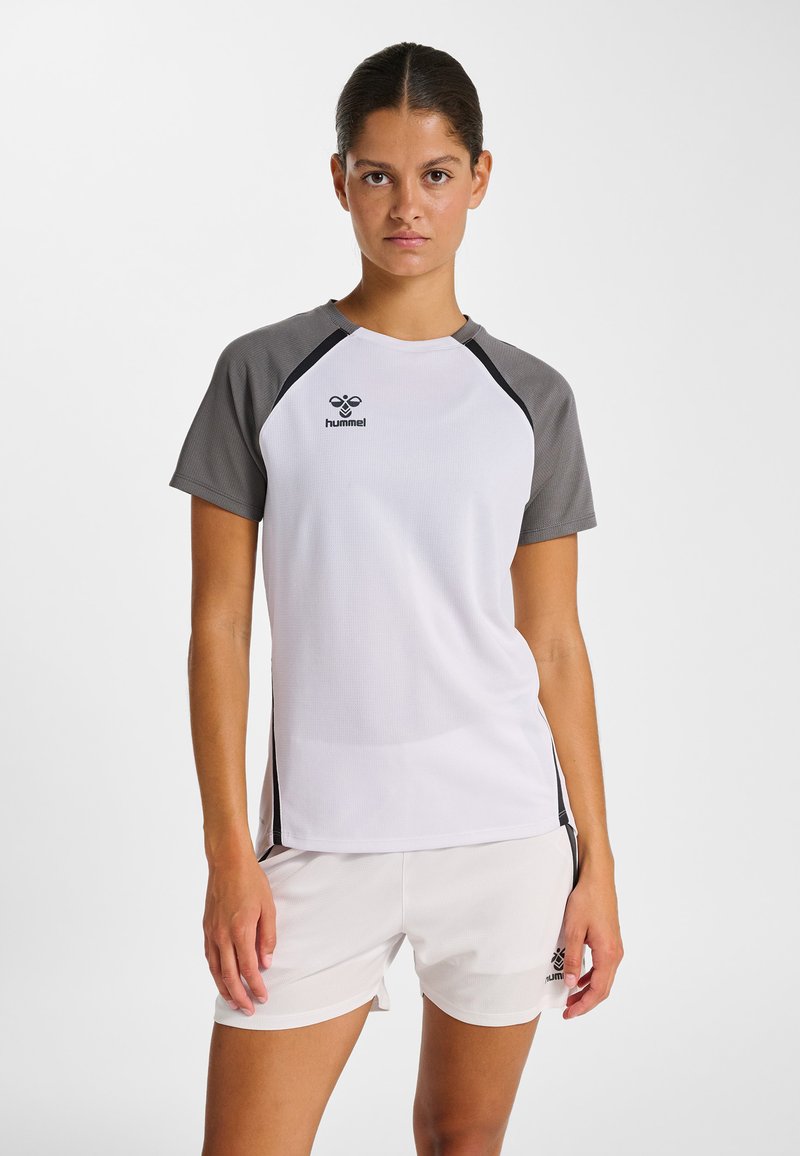 Femme debout portant un t-shirt de sport blanc avec des manches grises et un short de sport blanc, faisant face à l'avant sur un fond uni.