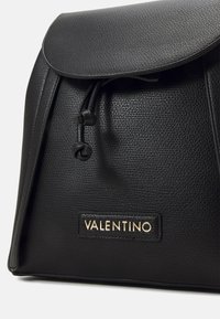 Czarna skórzana torba o fakturowanej powierzchni, z klapką z zakładką, regulowanym zamknięciem na sznurek i złotą plakietką z logo z przodu.