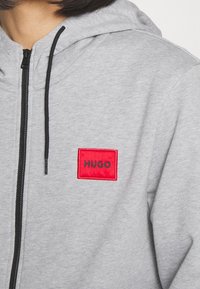 Sudadera con capucha gris con un parche de logo "HUGO" bordado en rojo. El material es una mezcla suave de algodón con cierre de cremallera y cordón negro.