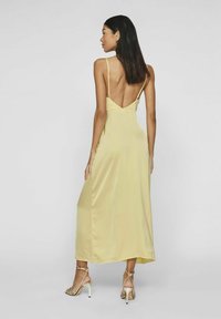 Vestido maxi de satén amarillo con tirantes finos y un escote bajo, con una textura suave y un diseño fluido. Combinado con zapatos de tacón con tiras.
