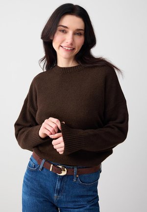 Jonge vrouw die glimlacht, gekleed in een donkerbruine trui en blauwe spijkerbroek met een bruine riem, staand tegen een effen lichte achtergrond.