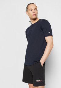 T-shirt in cotone blu navy con maniche corte, abbinata a pantaloni corti neri con logo "adidas ADVENTURE" in rosso e bianco, di una texture liscia.