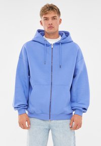 Felpa blu con zip realizzata in tessuto morbido, caratterizzata da un cappuccio con cordino, tasche a marsupio e polsini a costine. Indossati sotto un paio di jeans chiari.