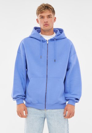 Felpa blu con zip realizzata in tessuto morbido, caratterizzata da un cappuccio con cordino, tasche a marsupio e polsini a costine. Indossati sotto un paio di jeans chiari.