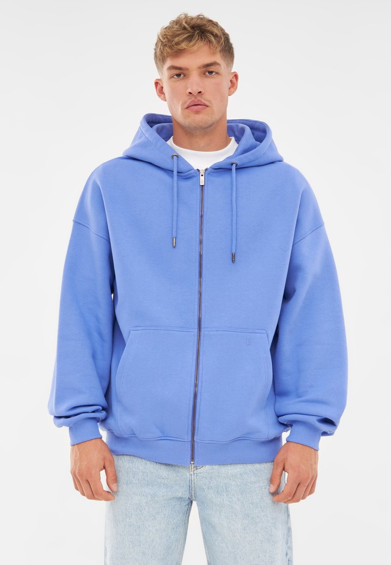 Sudadera azul con cremallera hecha de tela suave, con capucha ajustable, bolsillos tipo canguro y puños acanalados. Jeans de lavado claro debajo.