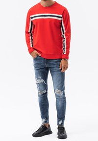Sudadera roja con rayas azul marino y blancas, combinada con jeans azules desgastados. Completada con zapatillas negras. Conjunto simple y casual.