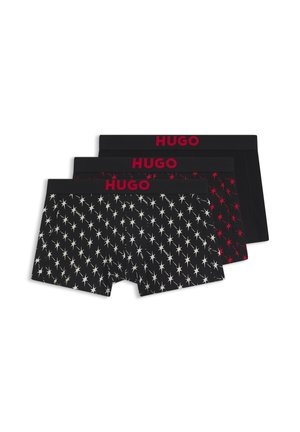 Jeune homme aux cheveux courts et foncés, torse nu, portant un boxer noir avec des motifs d'étoiles blanches et un logo rouge HUGO à la taille.