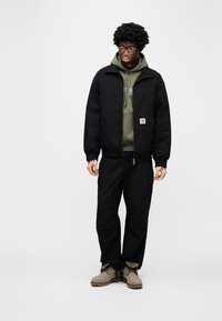 Μαύρο μπουφάν με φερμουάρ μπροστά, πράσινο φούτερ με κουκούλα από κάτω, μαύρο παντελόνι cargo και καφέ μπότες. Διαθέτει λογότυπο Carhartt στο μπουφάν.