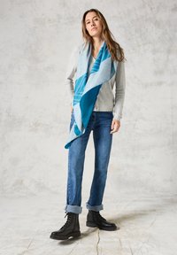 Blauwe en smaragdgroene sjaal met patroon van zacht materiaal, stijlvol gedrapeerd. Draagt men bij een grijze cardigan, blauwe jeans en zwarte veterschoenen.