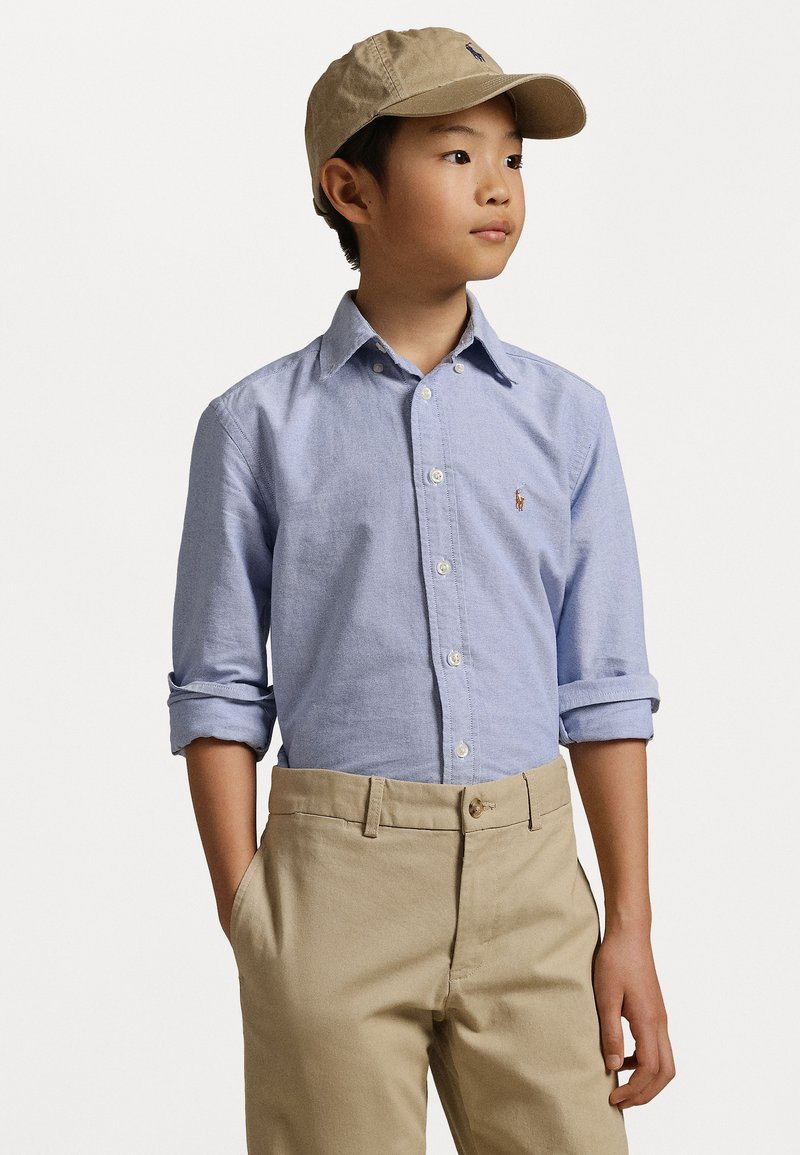 Ανοιχτό μπλε πουκάμισο με κουμπιά και σηκωμένα μανίκια, συνδυασμένο με μπεζ παντελόνι. Το πουκάμισο έχει ένα μικρό λογότυπο στο στήθος και κλασικό γιακά.