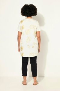 Tunique blanche avec motif floral doré, col rond et manches courtes, associée à un pantalon noir à la cheville avec un ourlet effiloché.