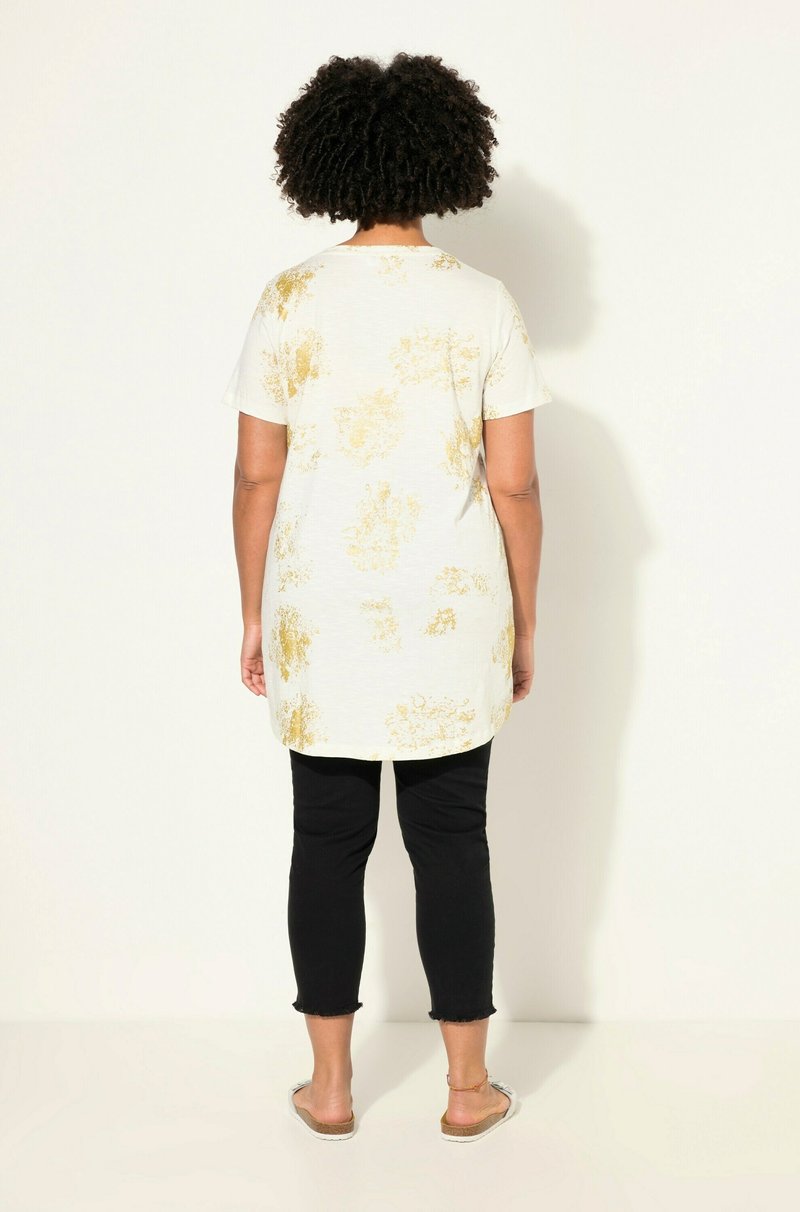 Tunique blanche avec motif floral doré, col rond et manches courtes, associée à un pantalon noir à la cheville avec un ourlet effiloché.