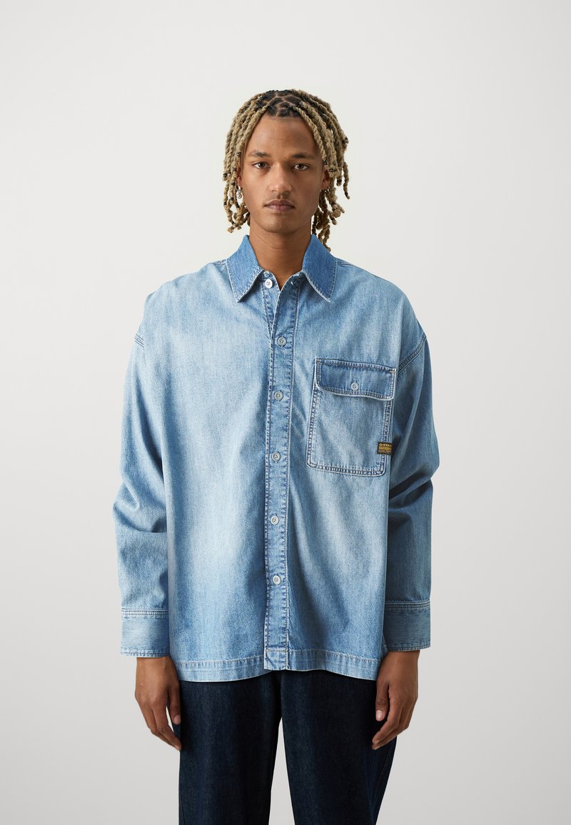 Světle modrá džínsová košile s button-up přední částí, dlouhými rukávy a kapsou na náprsní části. Má uvolněný střih a viditelné švy.