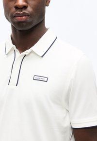 Homme portant un polo blanc Calvin Klein avec un bord marine sur le col, la patte de boutonnage et les bords des manches, visible des épaules au menton.