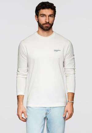 Homme aux cheveux bouclés foncés et à la barbe, portant une chemise blanche à manches longues avec un petit texte sur la poitrine et un jean bleu clair, debout devant un fond gris.