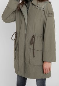 Olivgrön parkas med dragkedja och justerbar midja med dragsko, med infällda fickor och logotypdetalj på ärmen. Mjuk textur, avslappnad passform.