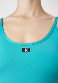 Débardeur turquoise avec une texture lisse, présentant un patch logo cK noir centré à l'avant et un col arrondi.