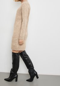 Robe-pull beige en maille avec un motif en zigzag texturé, longueur genou, poignets côtelés. Associée à des bottes cuissardes en cuir noir brillant.