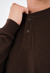 Cămașă lungă maro cu mânecă lungă, având un guler henley cu două butoane și un material texturat. Prim-plan al detaliilor gulerului și manșetelor.