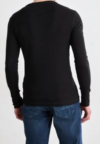 Mann in einem schwarzen, taillierten Langarmshirt und blauen Jeans, von hinten gegen einen schlichten hellen Hintergrund gezeigt.