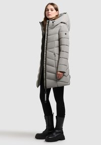 Ljusgrå pufferjacka med horisontell quiltning, överdimensionerad huva och två sidofickor med dragkedja. Stylad med svarta leggings och kraftiga svarta stövlar.
