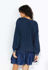 Blouse en bleu marine texturé à manches longues, avec un col froncé et des accents brodés sur les poignets, associé à une jupe en denim à volants.
