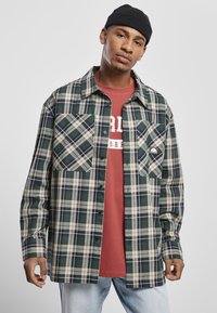 Mladi moški nosi črno kapo, zeleni in bež karo overshirt preko rdeče majice s potiskom ter svetlo modre jeans hlače na sivi podlagi.