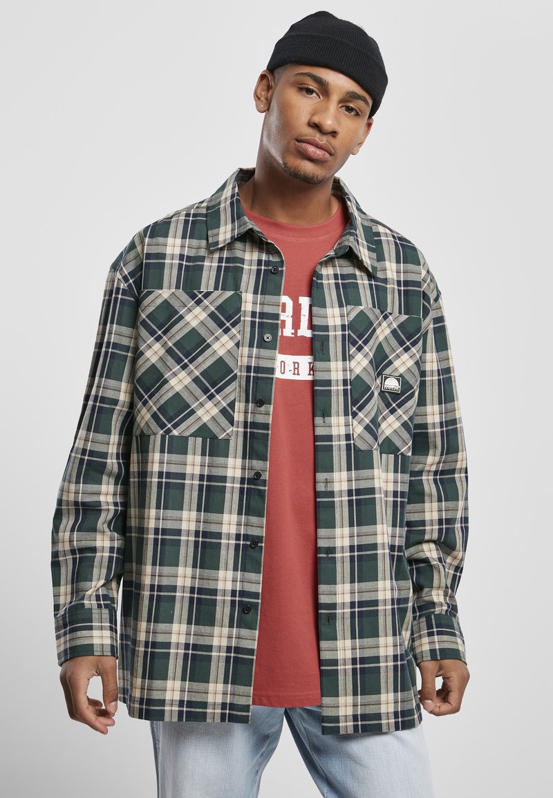 Mladi moški nosi črno kapo, zeleni in bež karo overshirt preko rdeče majice s potiskom ter svetlo modre jeans hlače na sivi podlagi.