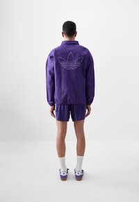 Lila jacka med elastisk nederdel och bakre Adidas-logotyp, i kombination med lila randiga shorts och vita strumpor, visar en sportig outfit.