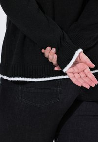 Maglione nero a maglia con polsini e orlo a coste bianchi, con texture. Jeans neri visibili, dettaglio della tasca posteriore in evidenza. Mani incrociate davanti.