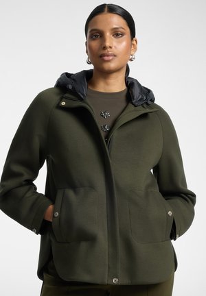 Jeune femme aux cheveux foncés portant une veste à capuche vert olive avec des poches devant, debout devant un fond clair uni.