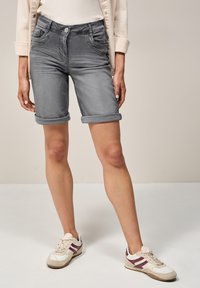 Pantalones cortos de mezclilla gris con dobladillos enrollados, diseño de cinco bolsillos y cierre de botón, combinados con zapatillas deportivas blancas y burdeos.