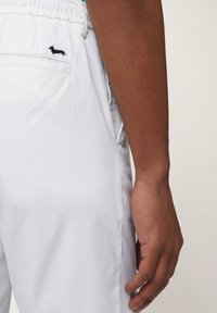 White cotton pants with an elastic waistband. Features a small navy embroidered logo on the back pocket, visible stitching, and a smooth texture.