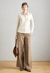 Beige Langarm-Hemd mit Knöpfen, kombiniert mit weit geschnittenen braunen Karohosen. Das Outfit umfasst eine braune Wildlederhandtasche und braune Absatzstiefel.