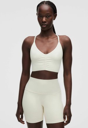 Woman with braided hair wearing light-colored fitted sports bra and high-waisted biker shorts standing against a plain background.