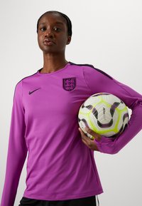 Hosszú ujjú, lila sportpóló fekete díszítéssel és jelvénnyel. A modell egy fehér és sárga labdarúgó labdát tart fekete részletekkel.