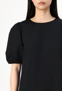 Svart t-skjorte med kort erm og rund halslinning og puffede ermer. Stoffet ser glatt ut med rynkede detaljer ved mansjetten.