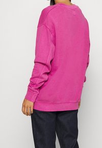 Sudadera rosa hecha de tela suave con un corte relajado, cuello redondo y puños acanalados; combinada con pantalones vaqueros oscuros.