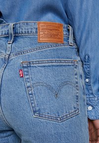 Blue denim jeans with a brown leather waistband label, featuring a red tab and iconic stitched pocket design. Light wash and textured fabric.