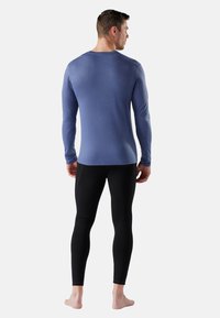 Man in een strak, blauw shirt met lange mouwen en zwarte leggings, staande op blote voeten, met zijn rug naar de camera gericht tegen een effen lichte achtergrond.