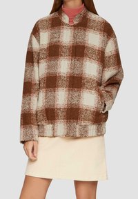 Brown and cream plaid oversized jacket with a high collar, textured fabric, side pockets, and dropped shoulders, worn over a light skirt.