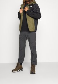 Chaqueta impermeable en verde oliva y negro, combinada con pantalones cargo grises con cremallera y botas de senderismo marrones. Detalle del logo reflectante.