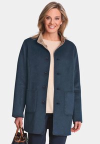 Marineblauer Wollmantel mit einem beigen Kragen, fünf schwarzen Knöpfen und zwei vorderen Taschen. Glatte Textur mit einer taillierten Passform.